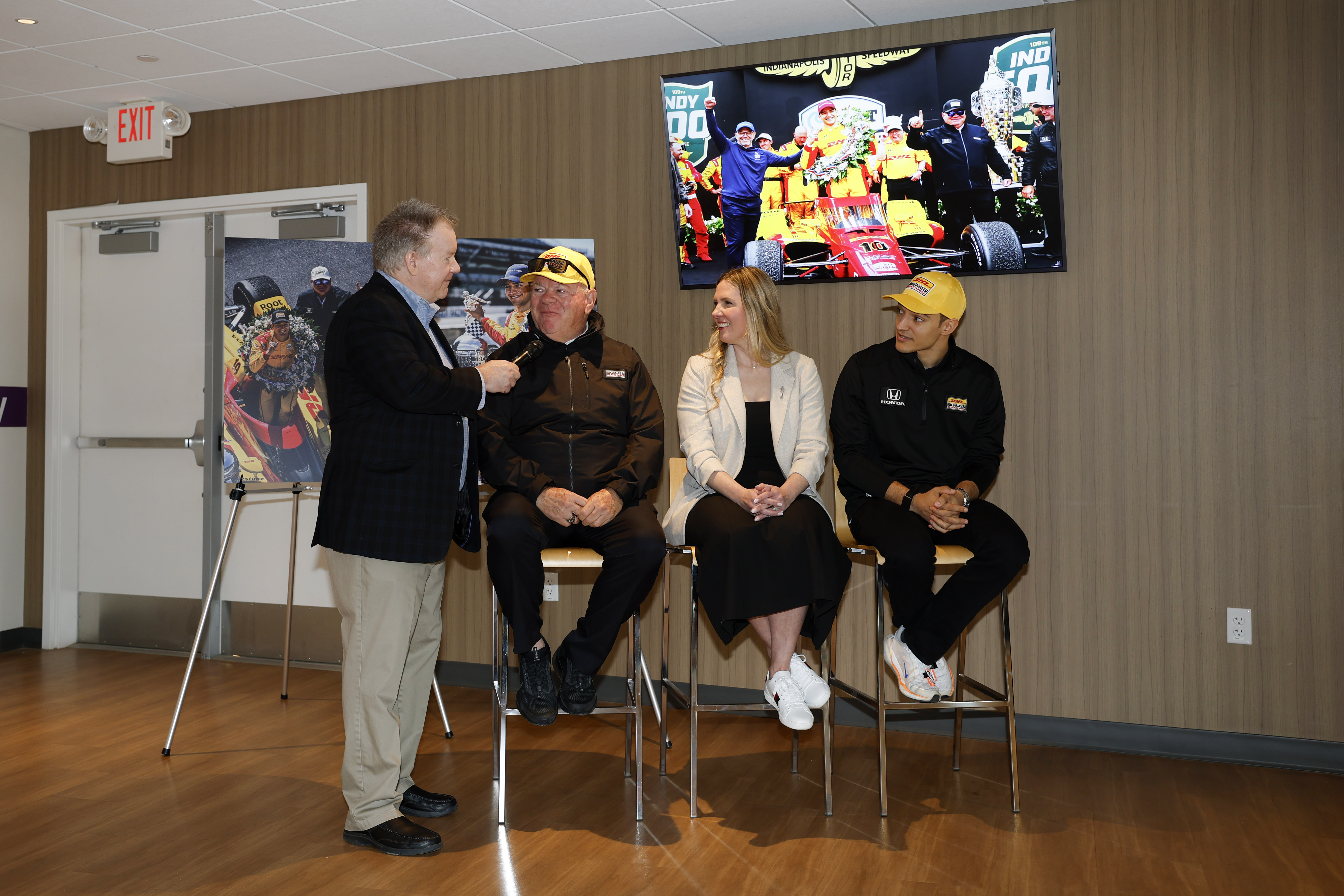 One man stands with a microphone and three others sit and smile