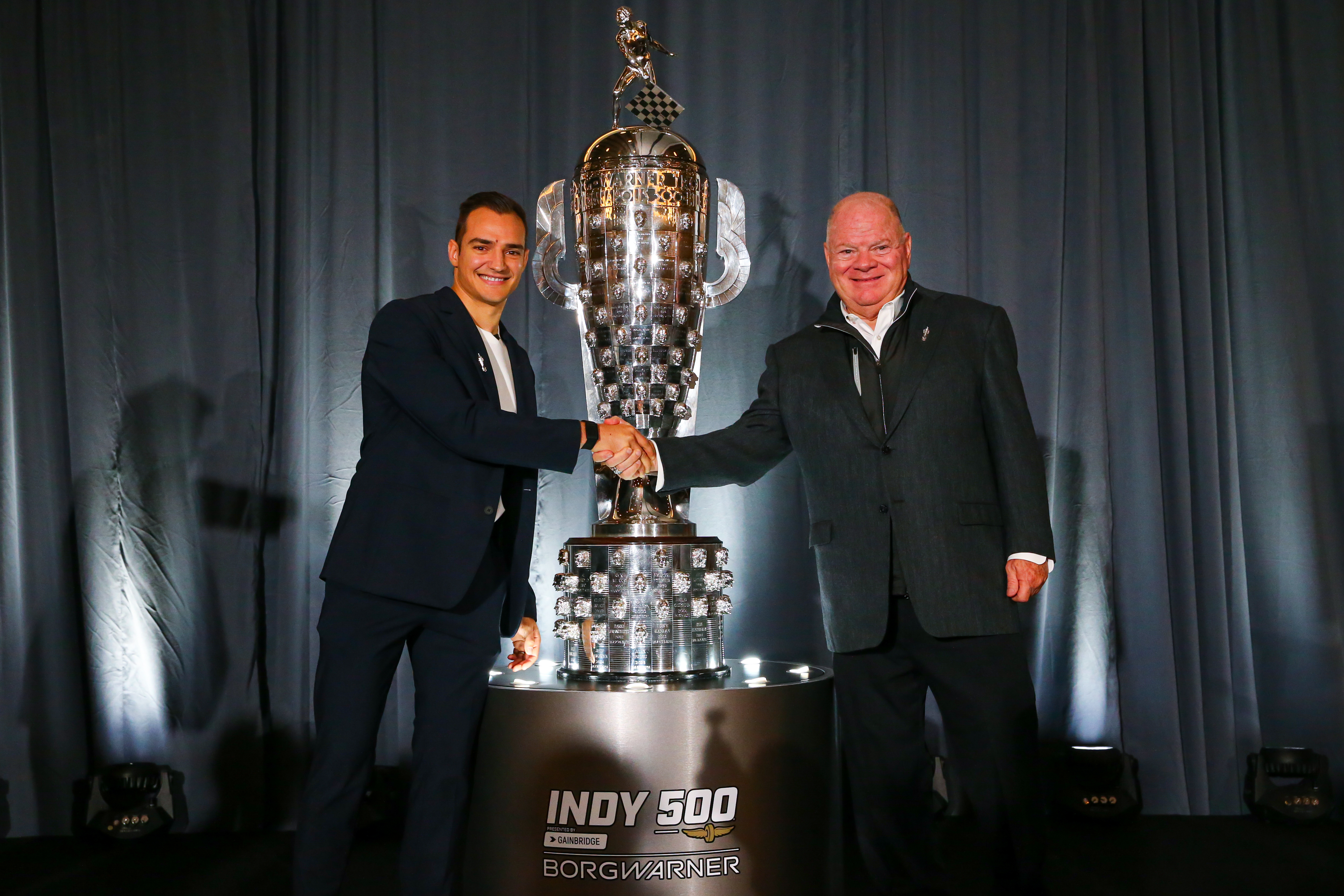 Two men shake hands onstage in front of large silver trophy