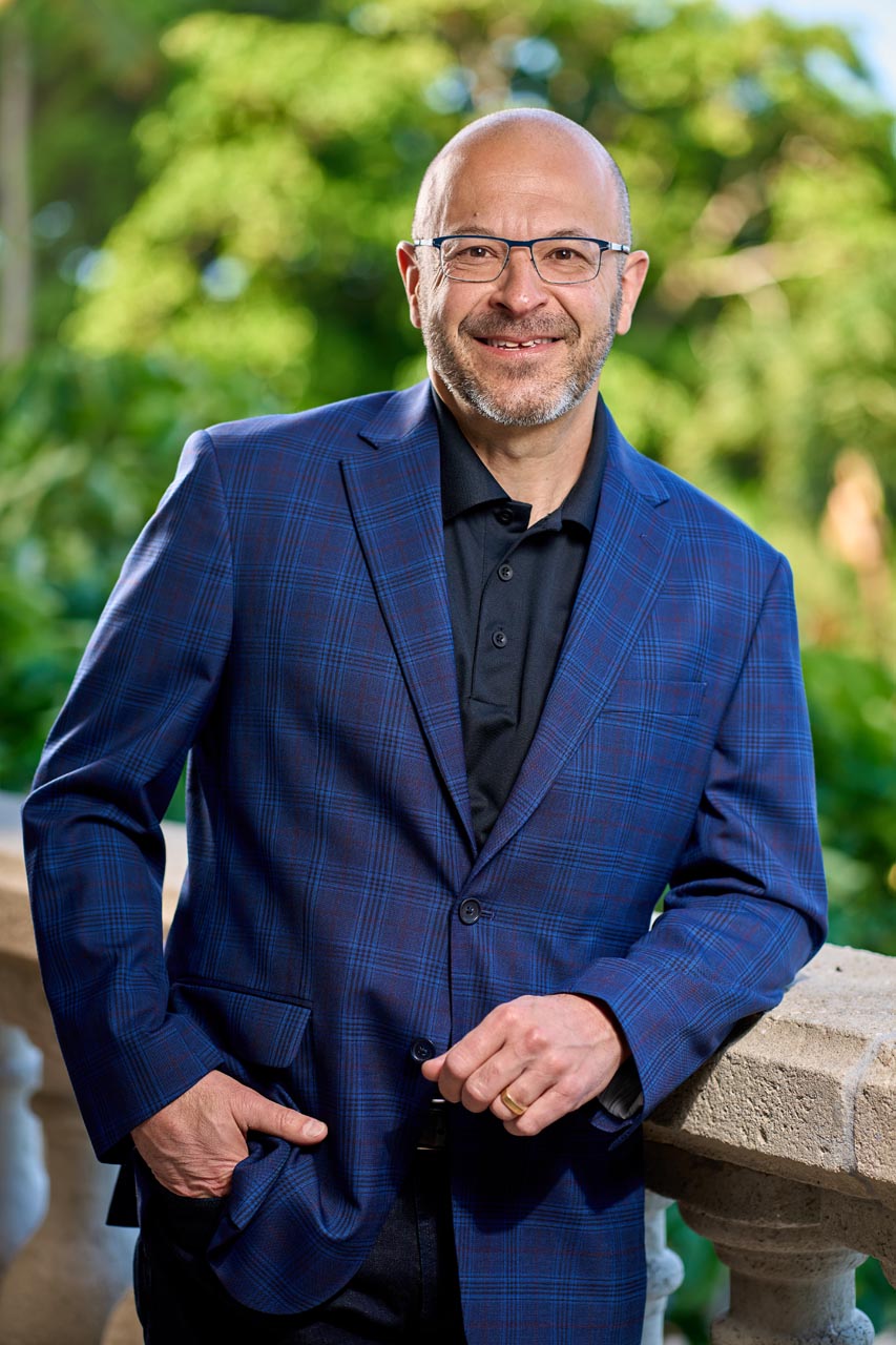 Smiling man in blue plaid suit leans against a bridge