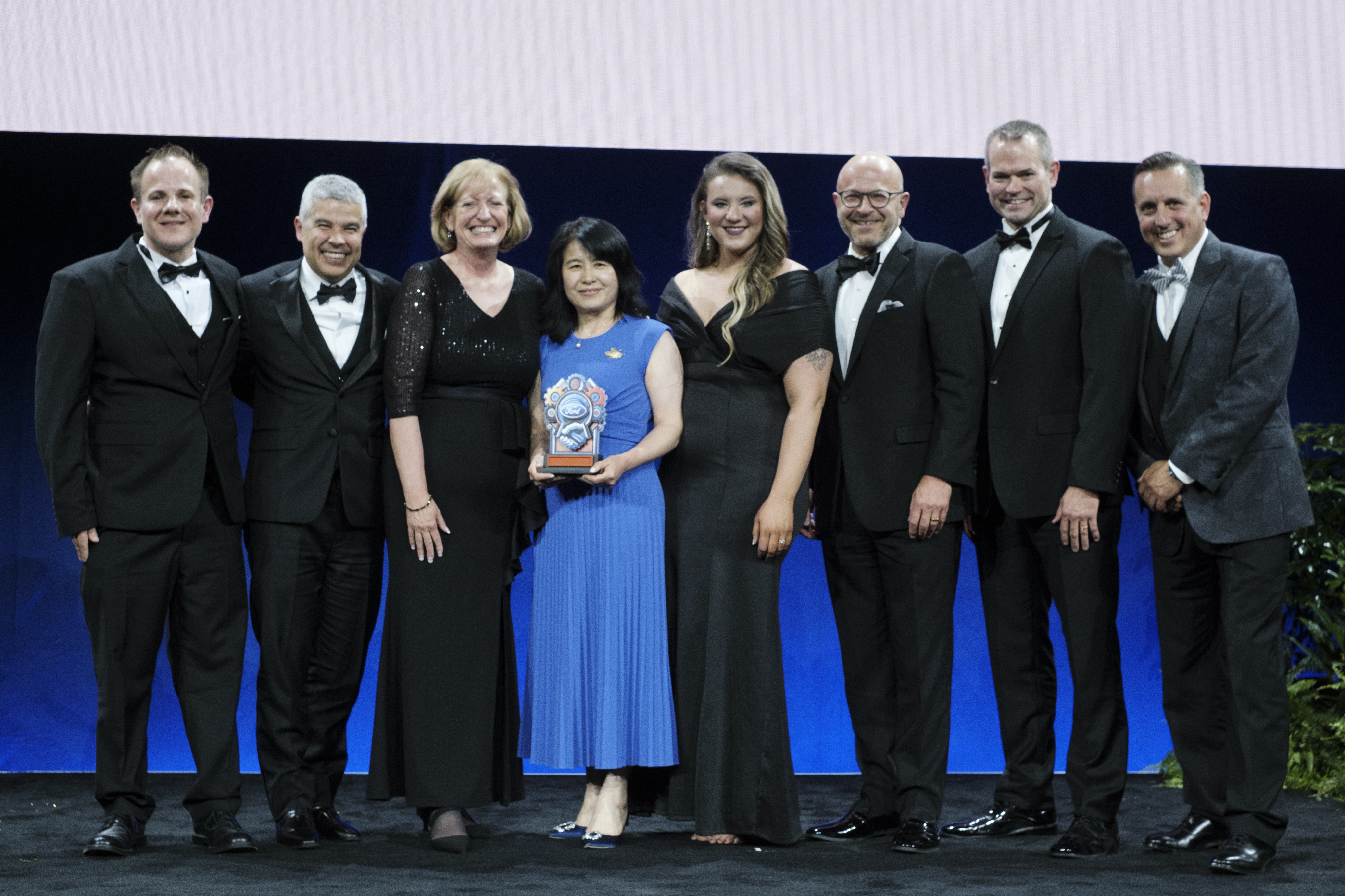 Group of 8 people in formal clothing stand on stage and hold trophy