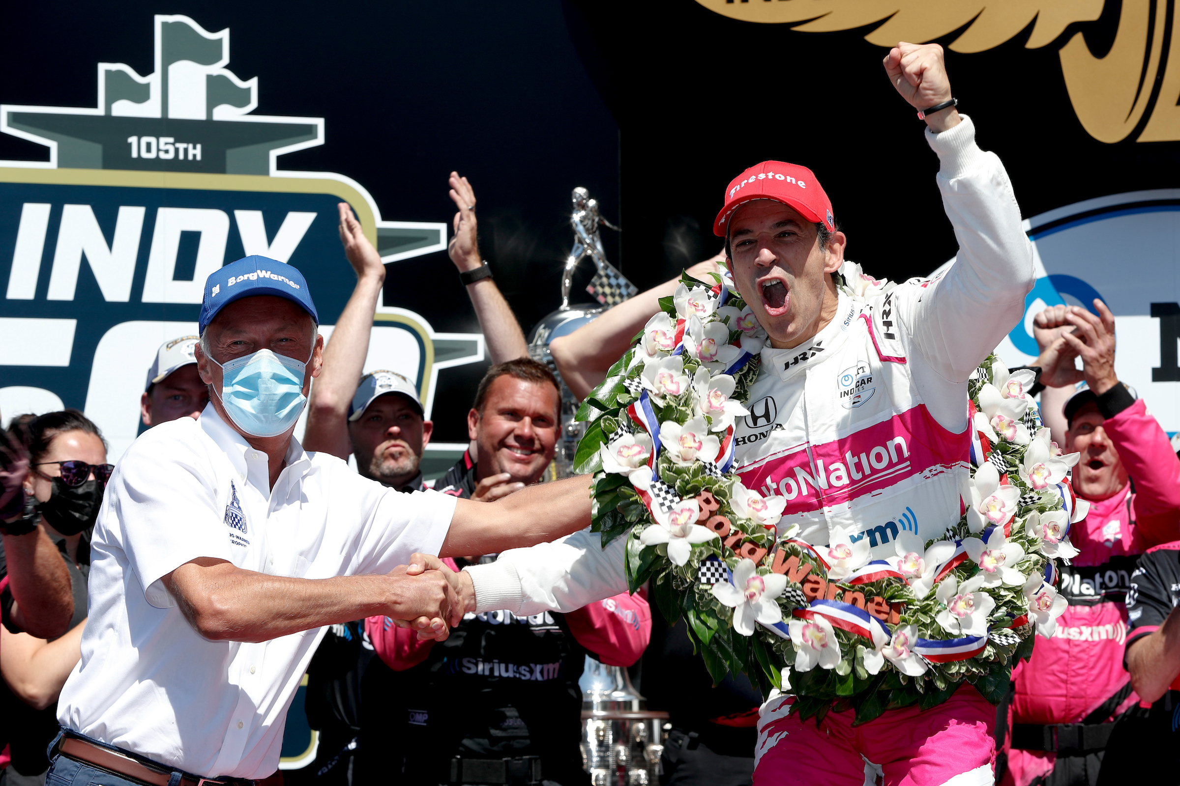 Man in blue hat shakes hand of man in pink hat with celebratory wreath with crowd in background