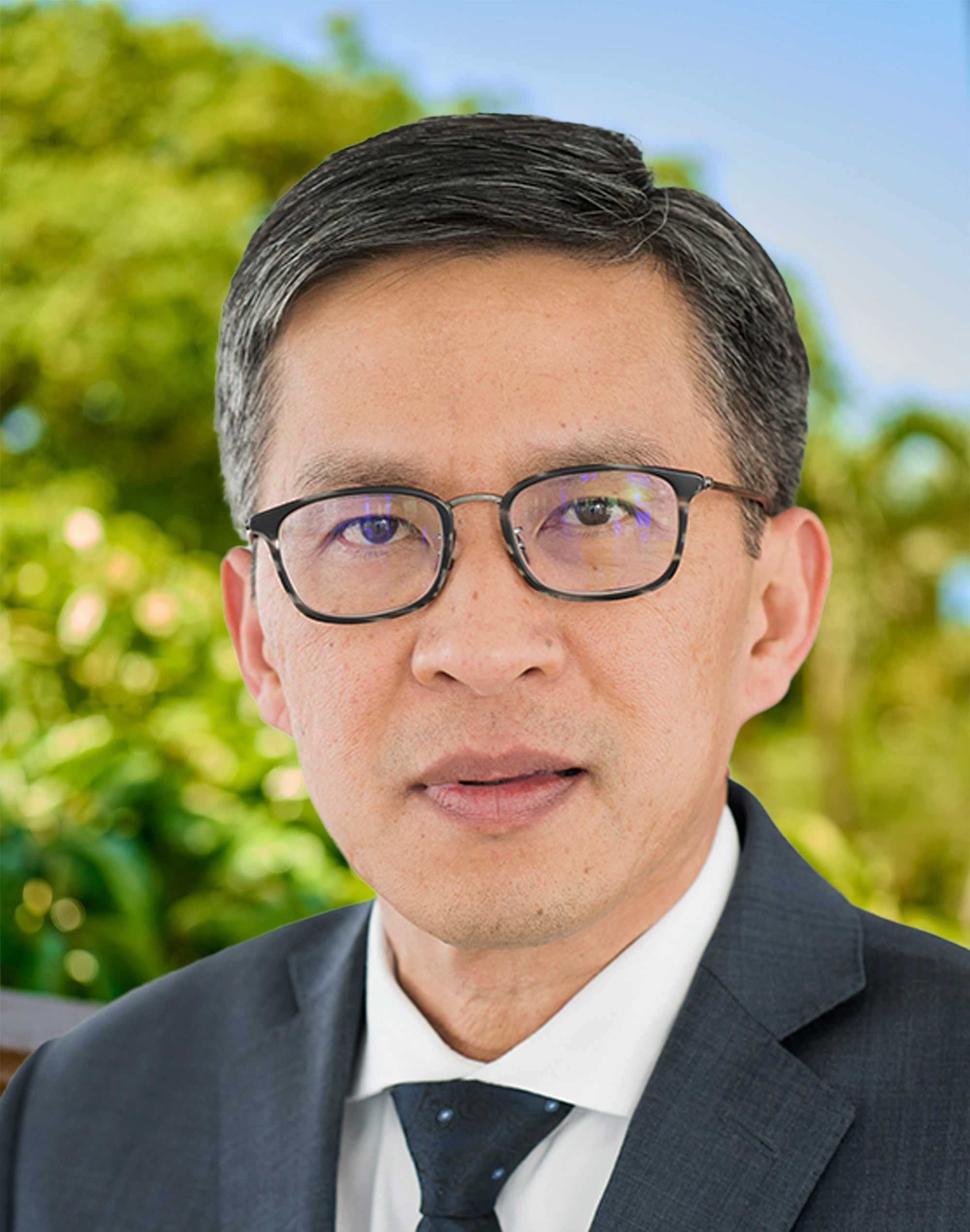 Man with glasses looks straight with blurred greenery in background