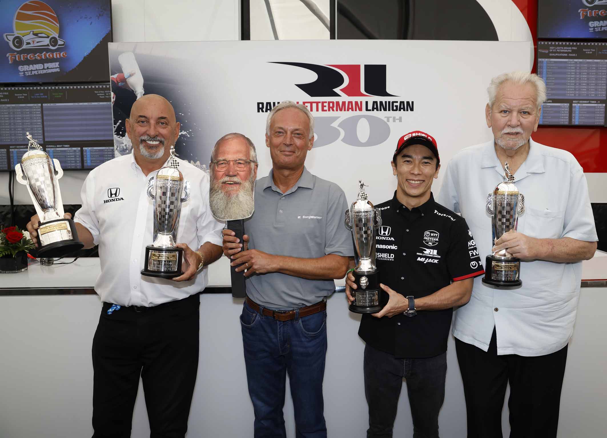 Group of four men stand in line holding trophies and head cut out of another man with a beard