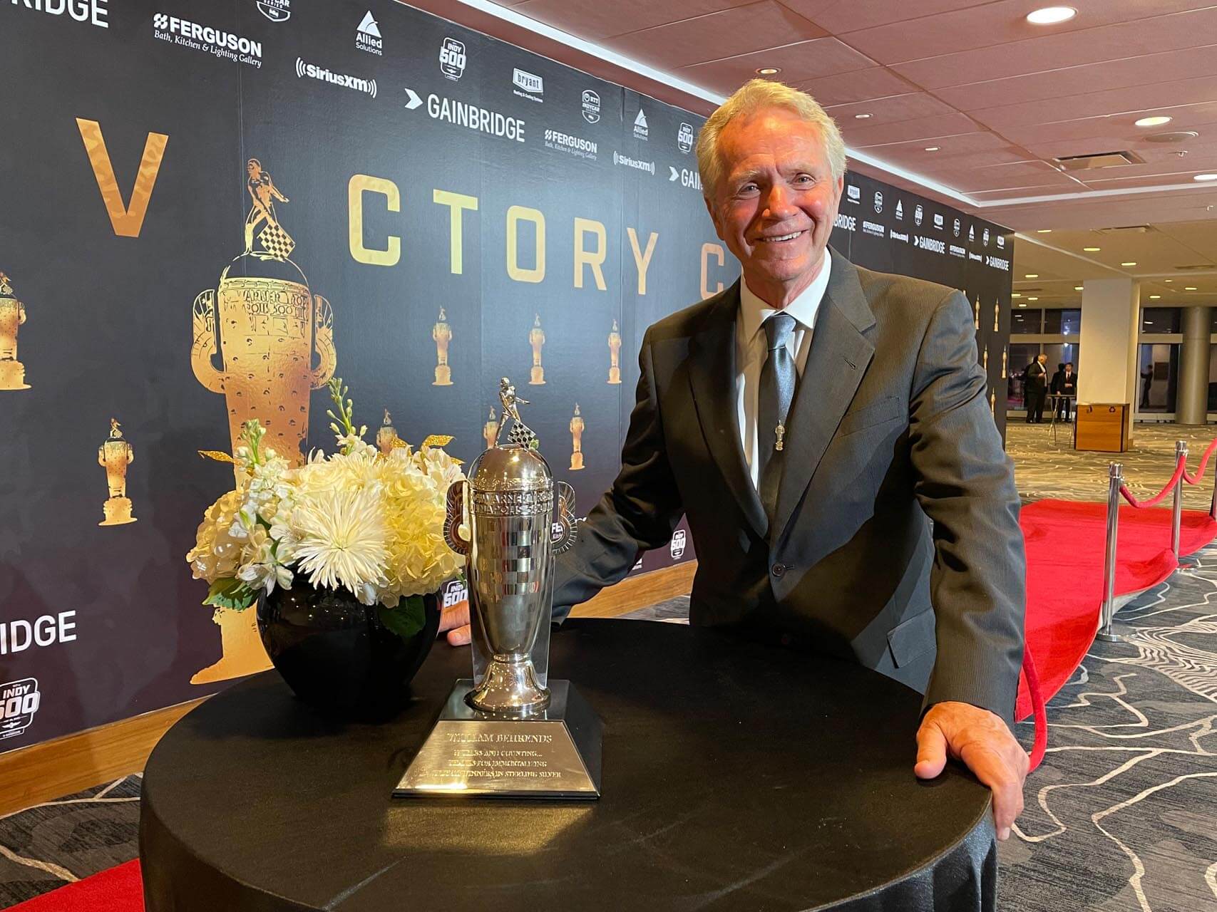 Man in suit jacket stands at table with white flowers and silver trophy