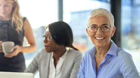 Who we are An experienced women with grey hair smiling at the camera while a team discusses in the back