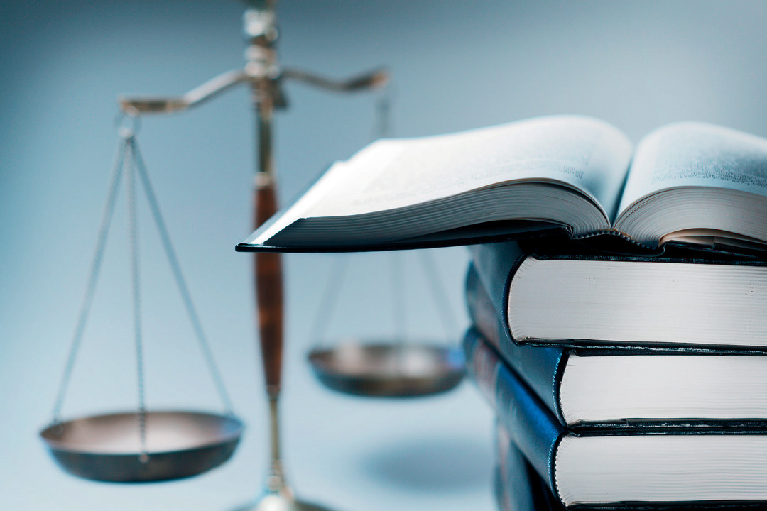 A stack of law books stands in front of a justice scale that is slightly out of focus.  On top of the stack is an open law book.